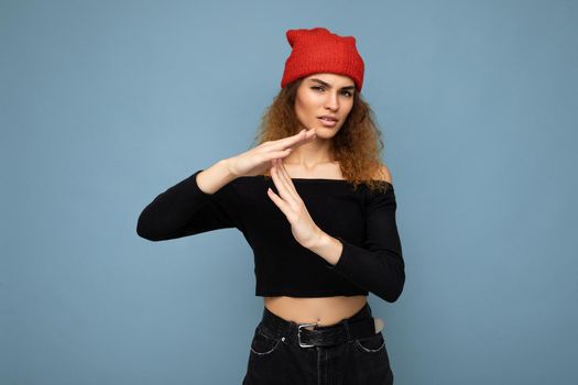 Young emotional dissatisfied pretty brunette woman wavy-haired with sincere emotions wearing black crop top and red hat isolated on blue background with free space and showing timeout gesture.