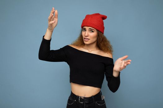 Young emotional dissatisfied pretty brunette woman wavy-haired with sincere emotions wearing black crop top and red hat isolated on blue background with free space. Don't understand concept.