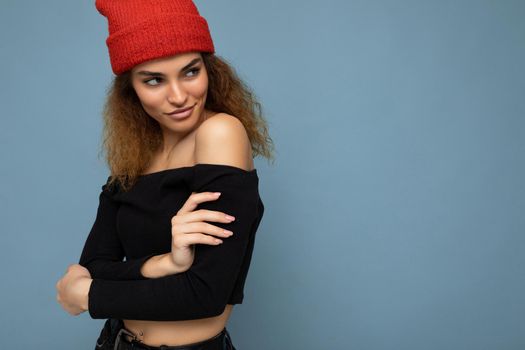 Photo of young positive sexy charming beautiful brunette woman curly with sincere emotions wearing stylish black crop top and red hat isolated on blue background with copy space and looking to the side.