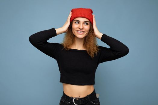 Photo of young positive smiling cute nice brunette woman curly with sincere emotions wearing stylish black crop top and red hat isolated on blue background with copy space.