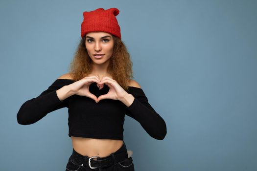 Photo of young positive cute nice brunette woman curly with sincere emotions wearing stylish black crop top and red hat isolated on blue background with copy space and making heart form with hands.