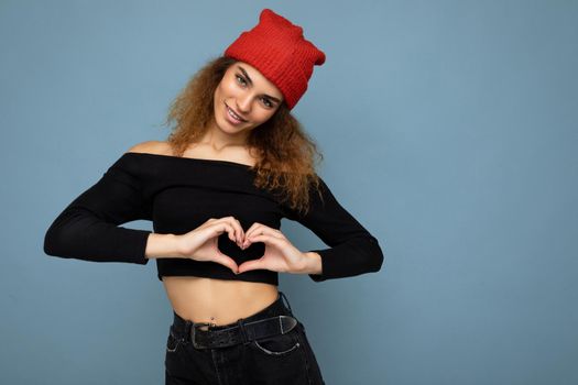Photo of young positive happy smiling beautiful woman with sincere emotions wearing stylish clothes isolated over background with copy space and showing heart gesture.