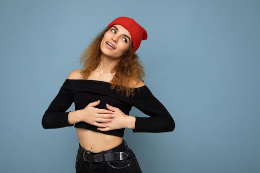 Photo of young positive cute nice brunette woman curly with sincere emotions wearing stylish black crop top and red hat isolated on blue background with copy space and holding hands on heart.