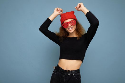 Photo of pretty beautiful attractive cool young curly dark blonde woman standing isolated over blue background wall with copy space for text wearing everyday trendy and stylish black top and red hat and glasses. Dancing and having fun.