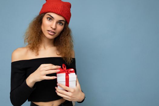 Shot of charming positive amazed young dark blond curly woman isolated over blue background wall wearing black crop top and red hat holding gift box looking at camera. Copy space, mockup