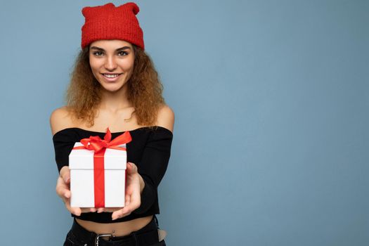 Beautiful happy young brunette woman isolated over colourful background wall wearing stylish casual clothes holding gift box and looking at camera. Empty space