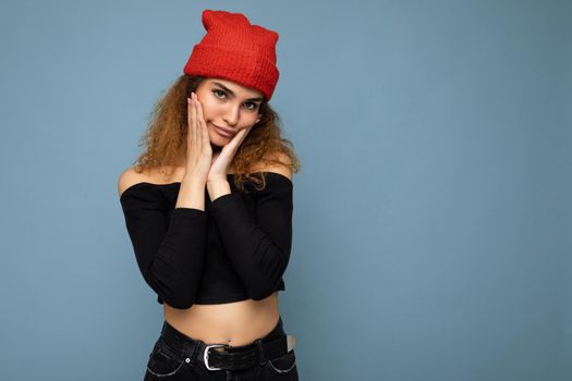 Photo of young positive funny cute nice brunette woman curly with sincere emotions wearing stylish black crop top and red hat isolated on blue background with copy space.