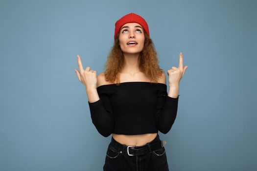 Photo of young surprised amazed attractive brunette curly woman with sincere emotions wearing black crop top and red hat isolated on blue background with empty space and pointing up at free space.