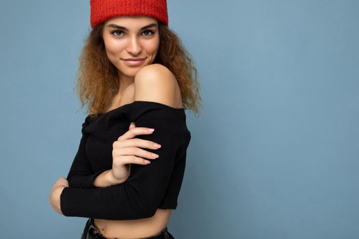 Photo of beautiful self-confident sexy young brunette curly woman isolated over blue background wall wearing black crop top and red hat looking at camera.