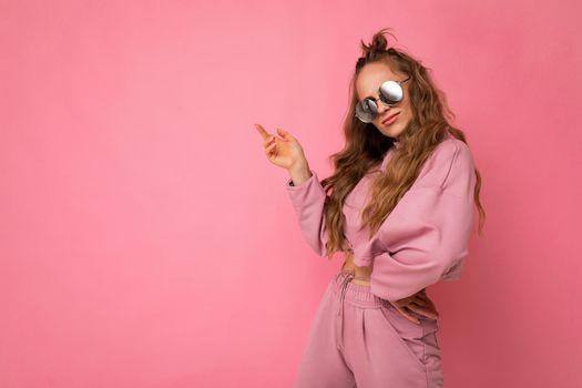 Photo shot of young beautiful attractive cute cool nice curly blonde woman wearing trendy and stylish pink sport suit and sunglasses isolated over pink background with copy space for text.