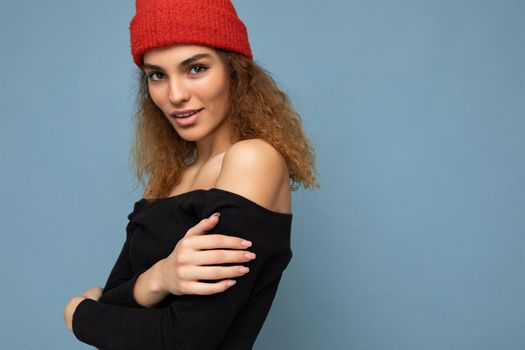 Close-up portrait photo of pretty beautiful attractive cool young curly dark blonde woman standing isolated over blue background wall with copy space for text wearing everyday trendy and stylish black top and red hat.