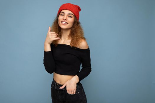 Photo of young positive cute nice brunette woman curly with sincere emotions wearing stylish black crop top and red hat isolated on blue background with copy space and imitating mobile phone.