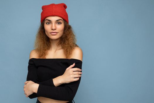 Photo of beautiful self-confident young brunette curly woman isolated over blue background wall wearing black crop top and red hat looking at camera. Free space, copy space