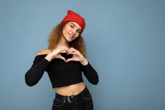 Photo of pretty beautiful attractive chatming smiling happy cool young curly dark blonde woman standing isolated over blue background wall with copy space for text wearing everyday trendy and stylish black top and red hat and showing heart and love gesture with hands.