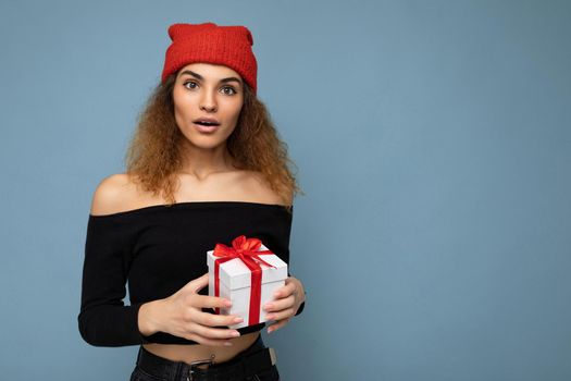 Photo shot of beautiful positive surprised young dark blonde curly woman isolated over blue background wall wearing black crop top and red hat holding gift box looking at camera. Copy space, mockup
