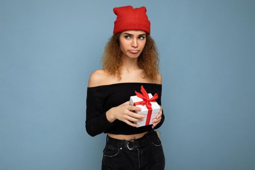 Photo of pretty beautiful attractive dissatisfied and unhappy young curly dark blonde woman standing isolated over blue background wall with copy space for text wearing everyday trendy and stylish black top and red hat. Holding white gift box with red ribbon.