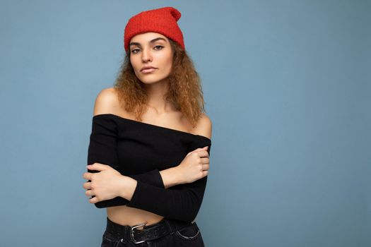 Photo of beautiful self-confident young brunette curly woman isolated over blue background wall wearing black crop top and red hat looking at camera. Copy space