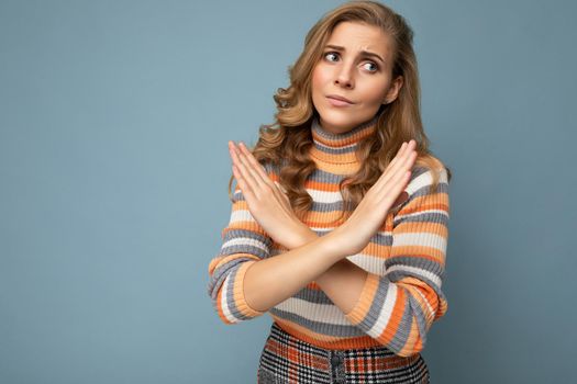 Portrait photo of young beautiful attractive dissatisfied unhappy blonde woman with wavy-hair wearing stylish striped sweater isolated over blue background with empty space for text and showing cross hands. No and stop gesture.