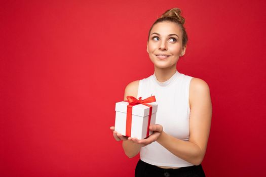 Photo shot of beautiful attractive cute positive dreaming smiling young blonde woman isolated over red background wall with copy space for text wearing casual white top holding gift box with red ribbon. Birthday concept.