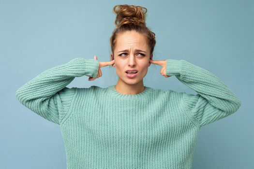 Portrait photo of young nice attractive pretty blond woman with sincere emotions wearing trendy blue sweater isolated over blue background with free space and covering ears.