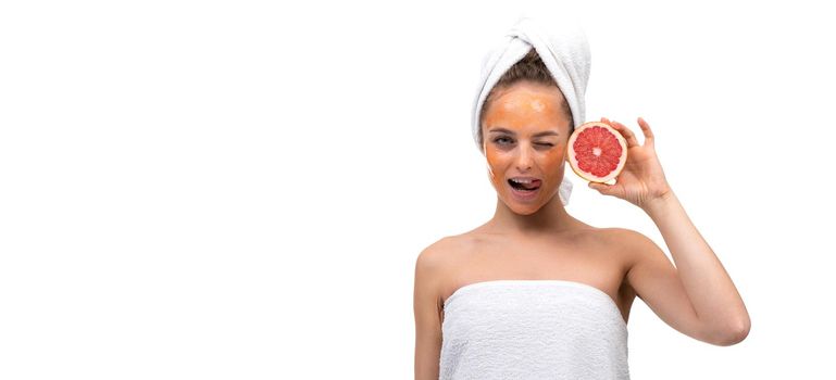 woman on a white background after a shower blinks one eye and holds a red grapefruit in her hands with a cosmetic mask on her face.