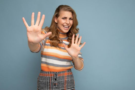 Portrait photo of young beautiful attractive dissatisfied and angry blonde woman with wavy-hair wearing stylish striped sweater isolated over blue background with empty space for text and showing no and stop gesture with hands.