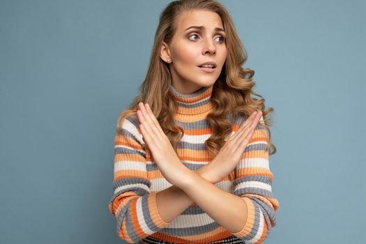 Portrait photo of young beautiful attractive dissatisfied unhappy blonde woman with wavy-hair wearing stylish striped sweater isolated over blue background with empty space for text and showing cross hands. No and stop gesture.
