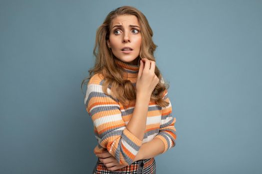 Portrait photo of young pretty beautiful attractive scared asking thinking blonde woman with wavy-hair wearing striped sweater isolated over blue background with empty space for text.