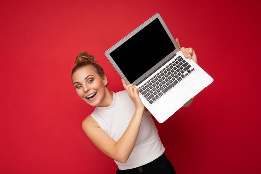 Photo shot of beautiful attractive positive happy smiling young blonde woman isolated over red background wall with empty space for text wearing casual white top and holding laptop computer with black blank display screen for mockup.