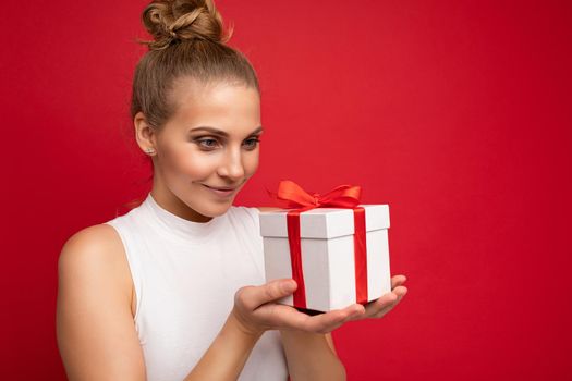 Photo shot of beautiful attractive cute positive smiling thinking young blonde woman isolated over red background wall with copy space for text wearing casual white top holding gift box with red ribbon. New year concept.