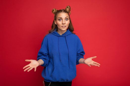 Photo of young charming beautiful attractive asking blonde woman with two horns with sincere emotions wearing stylish hipster blue hoodie isolated over red background with copy space and having questions.