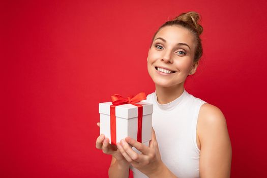Photo shot of beautiful attractive cute positive happu smiling young blonde woman isolated over red background wall with copy space for text wearing casual white top holding gift box with red ribbon. Surprise and present concept.