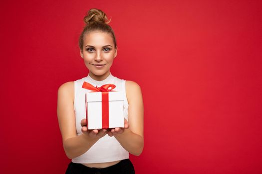 Photo shot of beautiful attractive cute positive young blonde woman isolated over red background wall with copy space for text wearing casual white top holding gift box with red ribbon. Birthday concept.