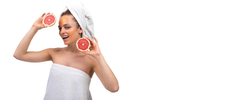 a woman after a shower with an orange cosmetic mask on her face is holding a cut grapefruit and dancing on a white background.