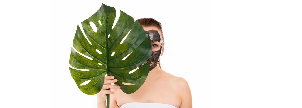 Concept of bath and spa procedures for body and face skin care of a woman in middle age, a young woman with a cosmetic black charcoal mask on her face and a palm leaf in her hands.
