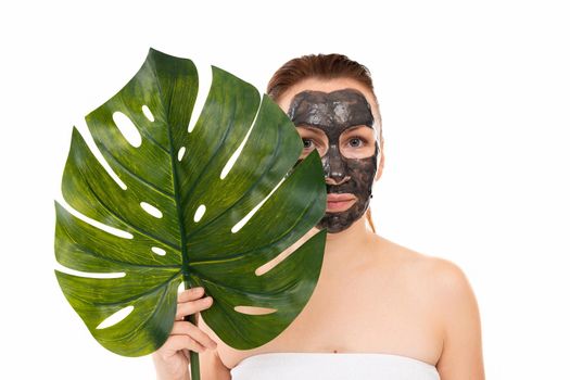 a woman in a black cosmetic mask on her face holds a huge palm leaf in her hands on a white background.