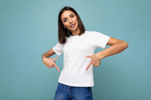 Photo of positive young fascinating beautiful attractive nice brunette woman with sincere emotions wearing casual white t-shirt for mockup isolated over blue background with copy space for text and fingers down.