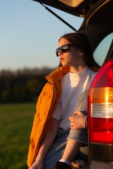 Pretty sad teenage girl with sun glasses sitting alone in a car trunk. Car travel concept.