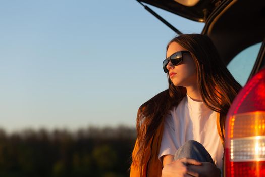 Pretty sad teenage girl with sun glasses sitting alone in a car trunk. Car travel concept.