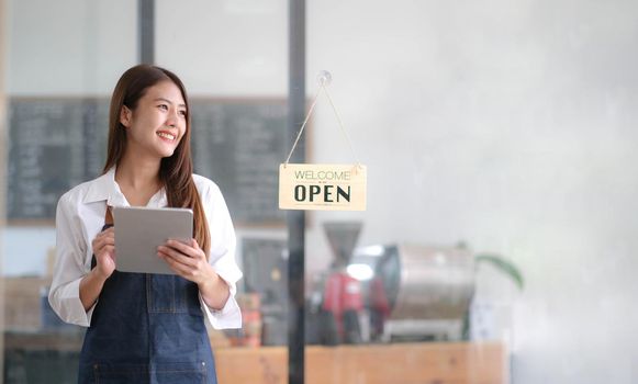 Asian Happy business woman is a waitress in an apron, the owner of the cafe stands at the door with a sign Open waiting for customers. Small business concept, cafes, and restaurants.