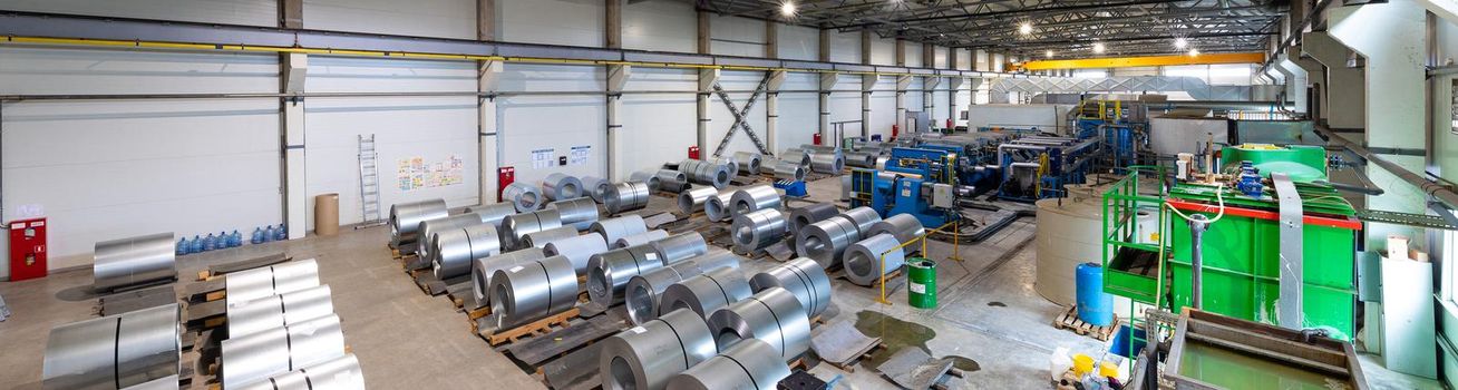 Panoramic photo shot of roll of painted galvanized steel sheet at cutting machine, ironworks and metalwork in factory. Industrial machine for metal sheet roof coils cut.