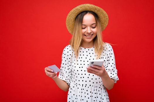 Photo of cheerful pretty blonde woman in summer outfit using mobile phone and holding credit card making payment online isolated over red background. Copy space.