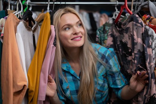 A young European woman sticks her head out from behind hangers full of clothes. The back room of a store and tailor's workshop. A blonde woman with straight long hair.