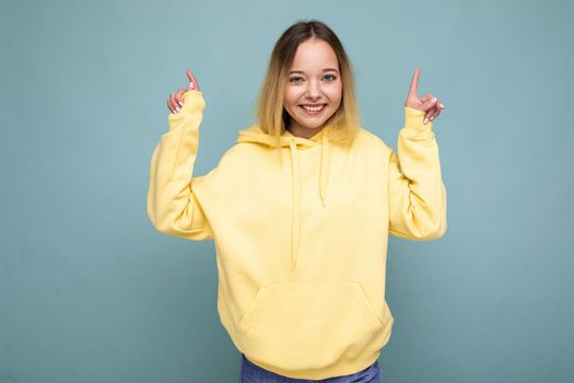 Young beautiful woman. Trendy woman in trendy yellow hipster hoodie. Positive female shows facial emotions. Funny model isolated on blue background with free space.