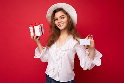 Portrait of positive cheerful fashionable woman in formalwear holding gift box and credit card looking at camera isolated on red background with copy space.