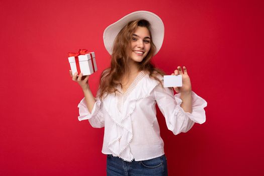 Young beautiful european stylish brunette woman wearing white blouse and fashionable hat isolated over red background with positive sincere emotions. Simple and natural looking at the camera. Holding gift box and credit card. Free space. Mock up.