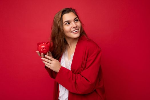 Young beautiful european stylish brunette woman wearing white blouse and red cardigan isolated over red background with positive sincere emotions. Holding little red moneybox. Free space.