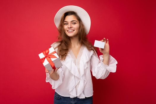 Portrait of positive cheerful fashionable woman in formalwear holding gift box and credit card looking at camera isolated on red background with copy space.