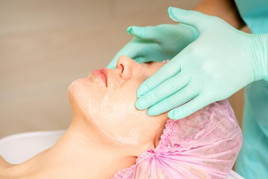 Cosmetologist with gloved hands applies a moisturizing mask with peeling cream on the female face. Facial cosmetology treatment. Procedures for facial care