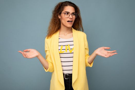 Photo of young dissatisfied astonished shocked beautiful brunette curly woman with sincere emotions wearing casual yellow jacket and optical glasses isolated on blue background with copy space and having doubts.